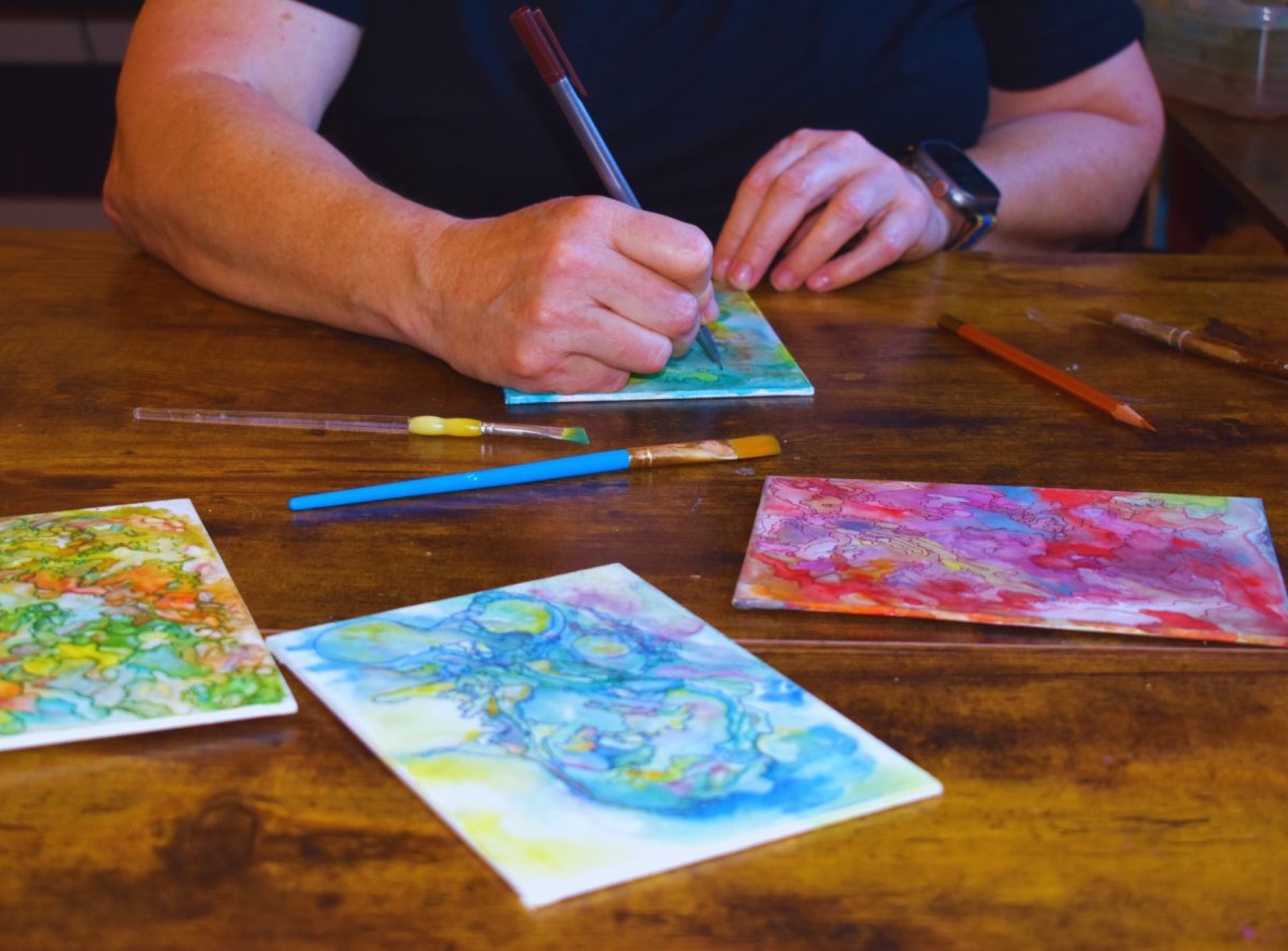 Diane's hands adding ink details to small watercolor cards at a wooden table